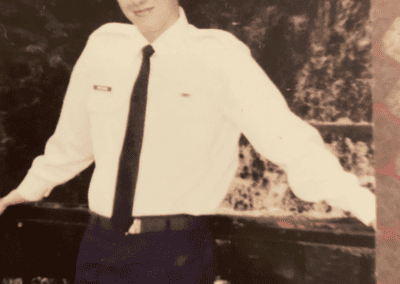 A young man in a white uniform shirt and dark trousers leans on a railing, smiling, representing a promotional product franchise.