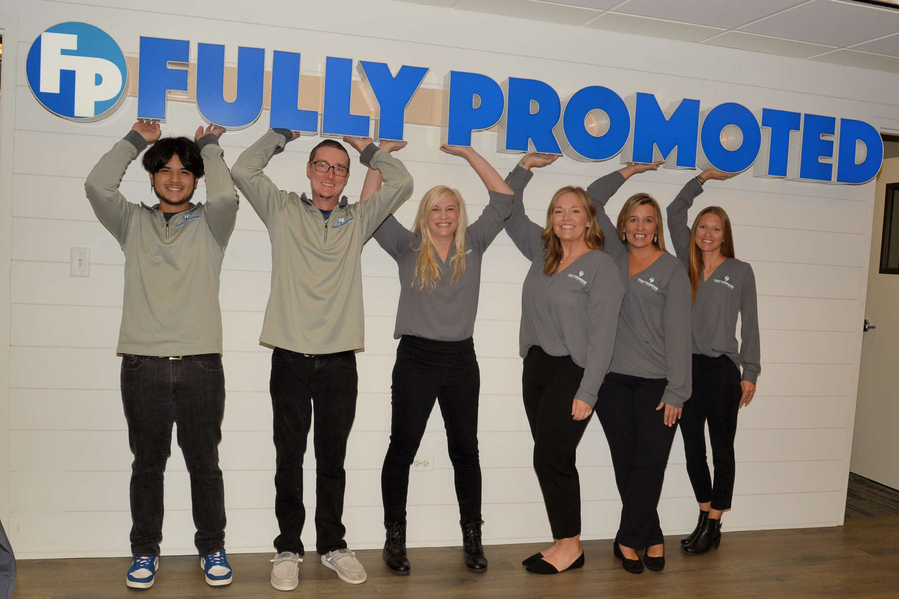 Five employees in gray shirts with the "fully promoted" branded apparel franchise logo pose with their arms raised in an office lobby.