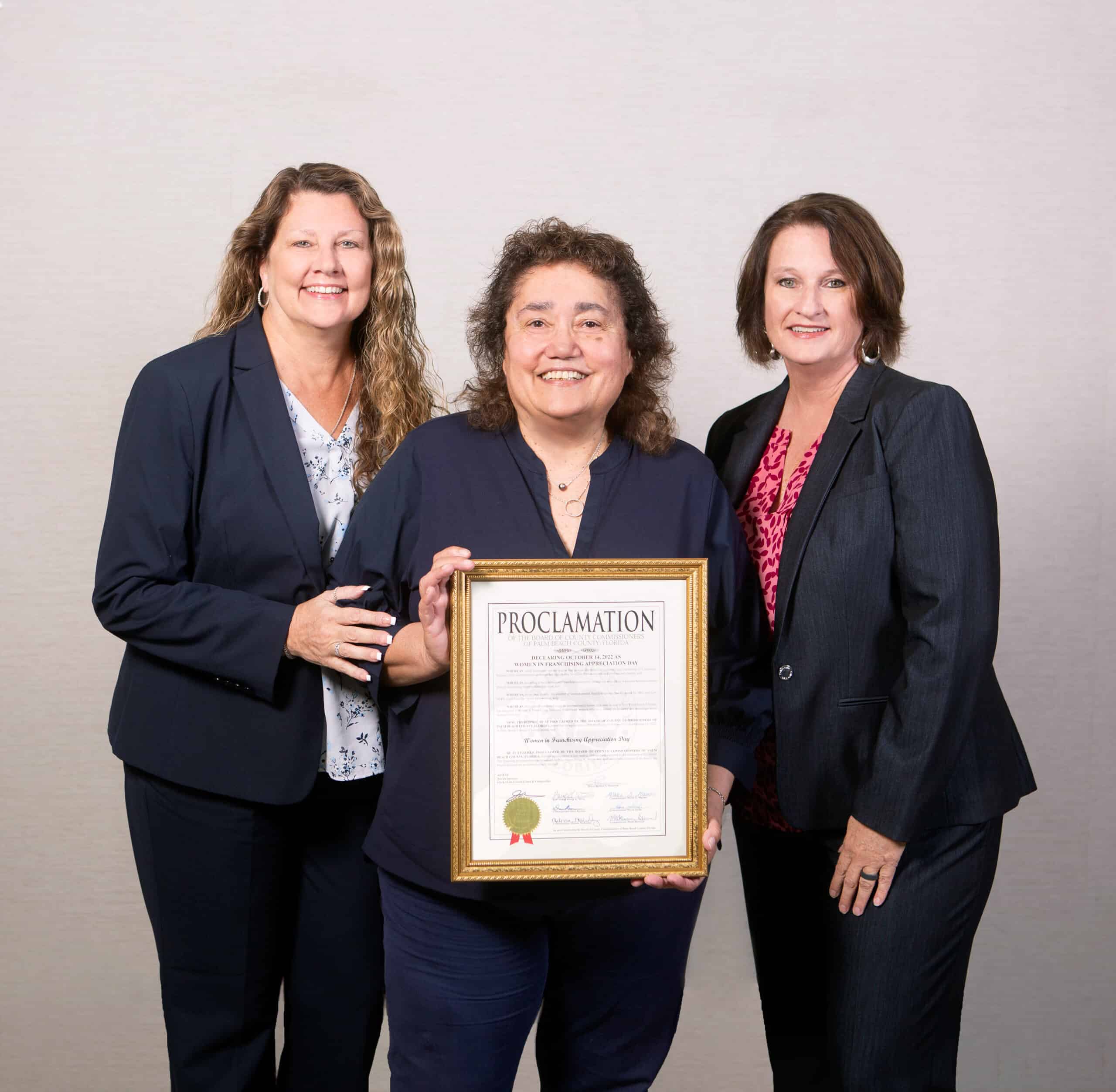 Three women smiling, standing together; one in the center holds a framed proclamation. They wear professional attire and are part of a branded apparel franchise.