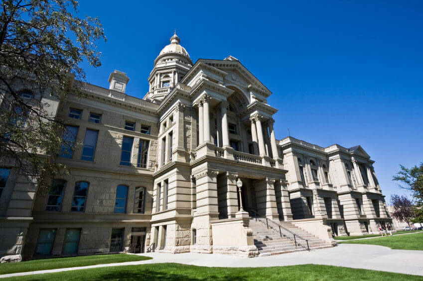 A large, historic government building with columns and a prominent dome, set against a clear blue sky with green lawns and trees in the foreground.