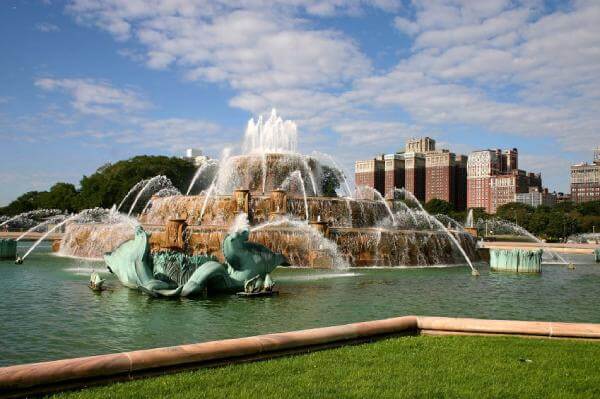 A large, ornate fountain with multiple water jets set in a park area surrounded by green grass; city buildings and a blue sky with clouds are visible in the background.