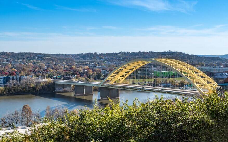 A yellow arch bridge spans across a river with cityscape and hills in the background under a clear blue sky. Vegetation is visible in the foreground.