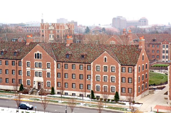 View of a large brick building with multiple windows, snow on the ground, and several other buildings visible in the background on a foggy day.