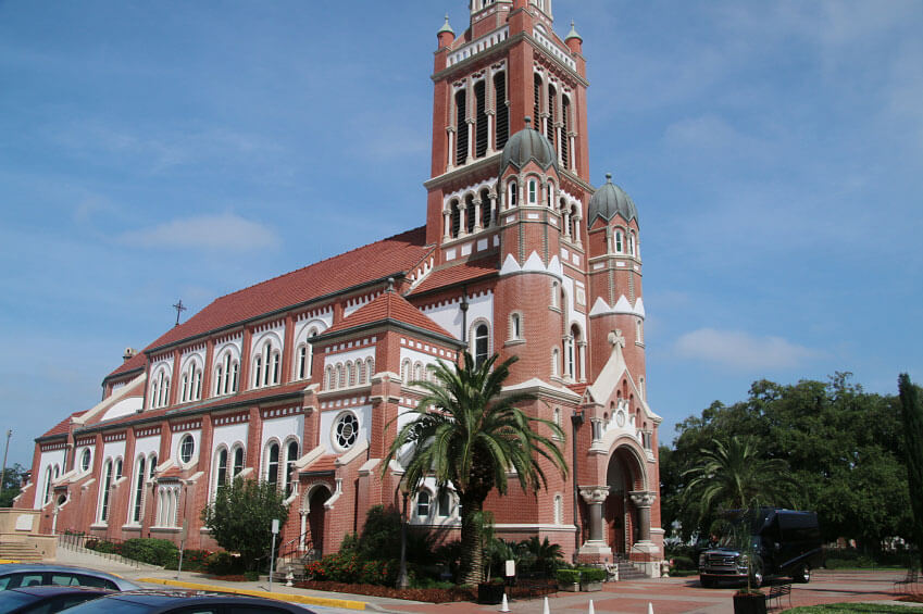 A red-brick church with a tall, ornate tower and arched windows, surrounded by palm trees and parked cars on a sunny day. A red-brick church with a tall, ornate tower and arched windows, surrounded by palm trees and parked cars on a sunny day.