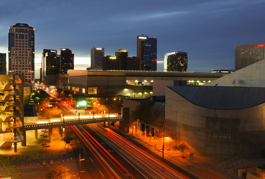 Nighttime cityscape featuring a mix of tall and low-rise buildings with illuminated windows, a road with light trails from moving vehicles, and a pedestrian bridge in the foreground.