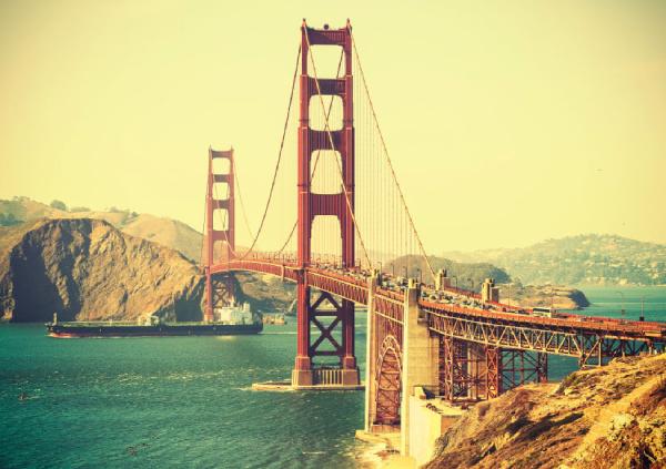 A large red suspension bridge spans a body of water, with hills in the background under a clear sky. A ship is passing under the bridge.