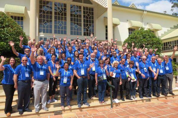 A large group of people wearing blue shirts stand on the steps of a building, smiling and waving at the camera.