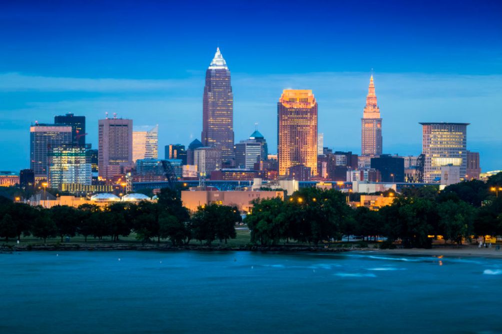 Skyline of Cleveland at dusk, featuring illuminated buildings against a blue sky, with Lake Erie in the foreground.