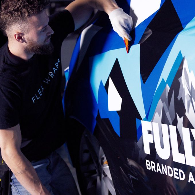 Person applying vinyl wrap on a car door with geometric blue and black design, wearing a black "Flex Wraps" t-shirt.