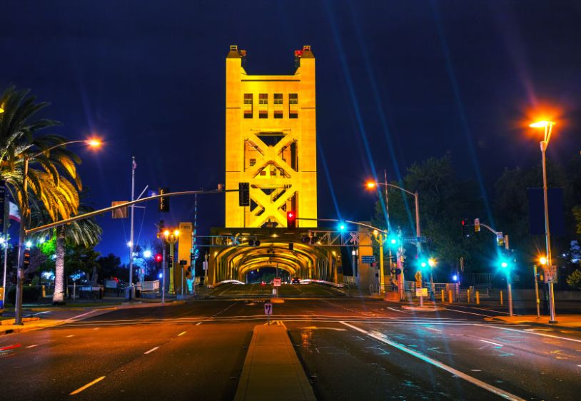 Night view of a brightly lit golden tower bridge with traffic lights and a palm tree on the left.
