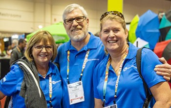 Three people wearing blue shirts and name badges smile at a convention. Colorful umbrellas are visible in the background. Three people wearing blue shirts and name badges smile at a convention. Colorful umbrellas are visible in the background.
