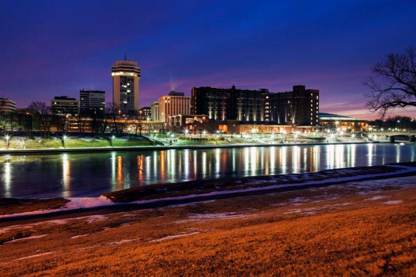 Cityscape at dusk with illuminated buildings reflecting on a calm river, surrounded by grass and bare trees under a deep blue and purple sky.